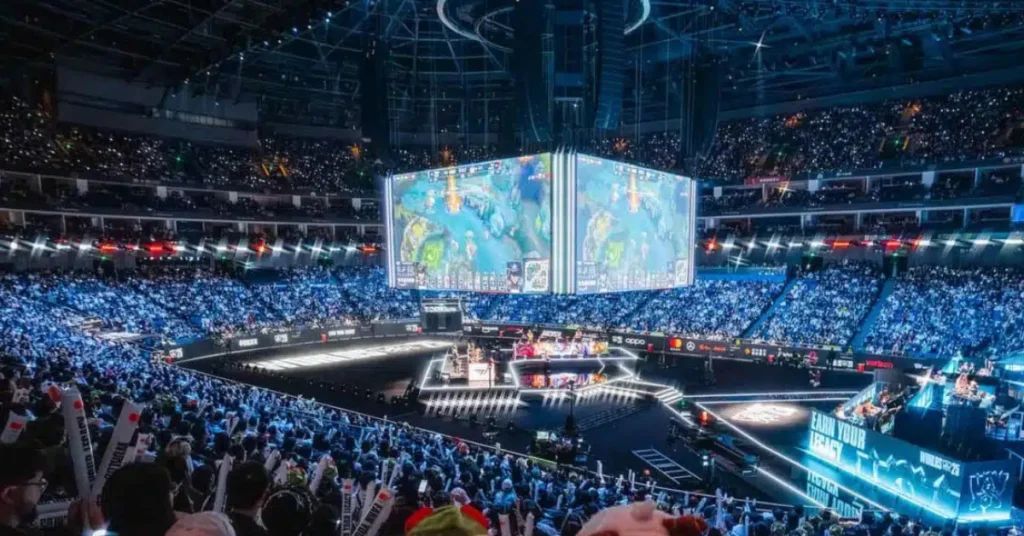 Massive esports stadium with four-sided screen showing MOBA gameplay, teams competing on stage, and cheering crowd.